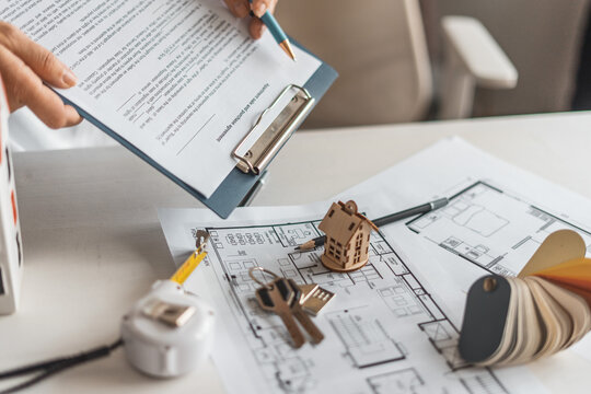 Hands holding property contract above house plans with keys, model home and color samples on desk. Real estate purchase, mortgage agreement and interior design planning concept for buyers.