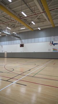 Wide shot of a clean, empty indoor gymnasium. Features a shiny wooden floor with colorful painted lines, a portable basketball hoop, and a high ceiling.