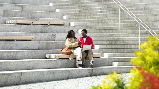 African american man and woman friends discussing news or work while sitting on stairs outdoors, positive male and female students buddies talking, sharing interesting information and thoughts.