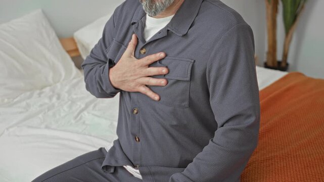 Man grey haired with beard clutching chest with hand while seated on bed wearing pajamas beside white sheets and orange blanket in building; pain health concern.
