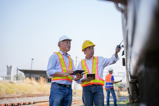 Engineers using digital tablets and walkie-talkie to inspect freight train at rail yard, Professional rail inspectors and technical team discussing logistics and maintenance near cargo train