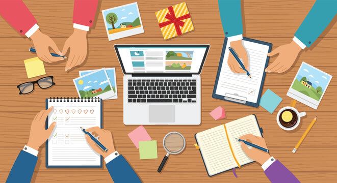 Top view of a collaborative team working at a wooden desk with a laptop, notebooks, printed photographs, and colorful sticky notes for planning.