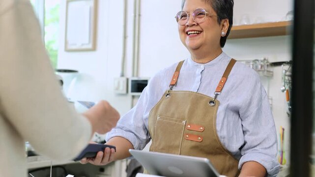 Senior entrepreneur cafe owner barista processes credit card payment on terminal, Asian female in apron smiling toward client across tiled counter, secure cashless sales inside modern coffee shop