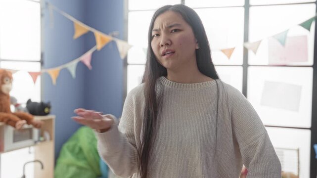 Young woman gesturing with open hand beside stuffed monkey and colorful bunting in a classroom play area, interacting with toys; mild frustration.