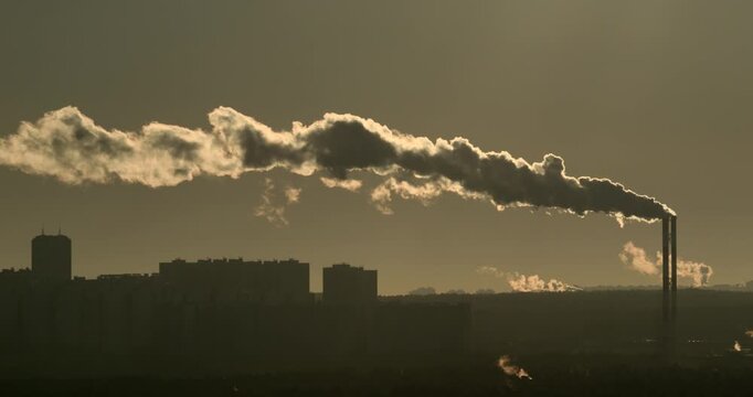 Tall industrial chimney releasing long dense plume above residential skyline in warm golden backlight Telephoto urban scene with haze district heating infrastructure emissions and air pollution mood