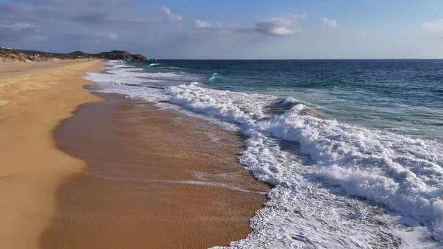 Drone flying low over crashing waves along the coast in Baja California Sur, following the shoreline with warm sunset light off to the side.