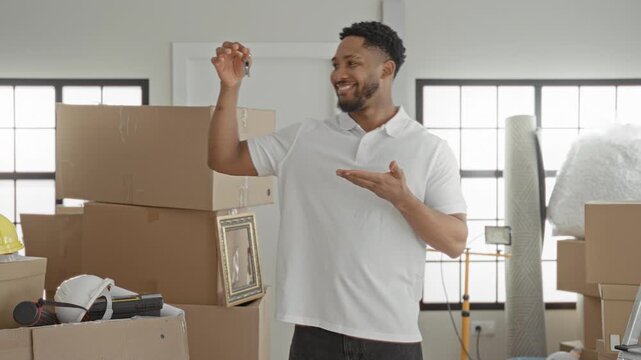 Man holding keys and smiling, gesturing with one hand beside stacked moving boxes and packing tape in building; new home joy.