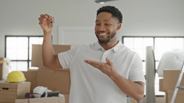 Man holding keys in raised right hand and presenting them with left palm toward stacked moving boxes, hardhat and ladder inside a bright building, smiling; new home joy.
