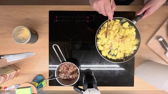 Top-down view of a person cooking scrambled eggs in a non-stick pan. The wooden counter holds various ingredients and kitchen tools. Bright lighting creates a cozy kitchen atmosphere.