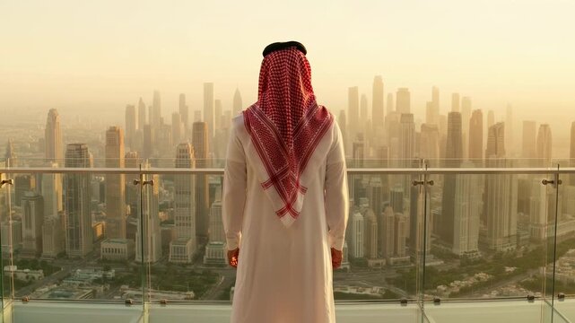 Man wearing white thobe and red keffiyeh stands on high-rise balcony gazing at sprawling cityscape below in golden morning light.