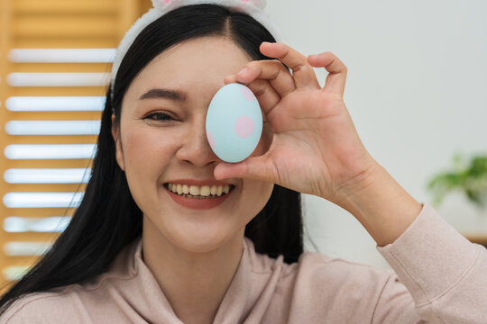 happy woman wearing bunny ears playing close her eyes with egg and laughing at home on easter day