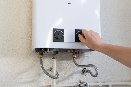 Person's hand turning a knob on a white wall-mounted gas water heater, demonstrating installation, maintenance, or repair work on heating equipment and home appliances