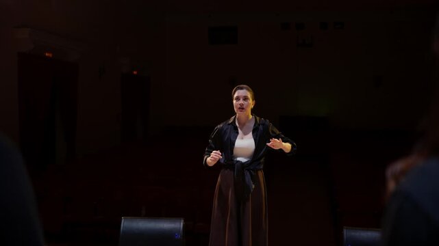 Interpreter on dimly lit stage uses expressive sign language and gestures to communicate with audience members during quiet performance rehearsal in empty theater