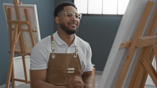 Young man in apron painting at studio and then leans back with hands behind head; creative contentment.