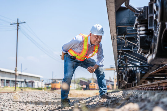 Senior industrial worker checking mechanical parts of a train undercarriage during daylight, Railway engineer in safety vest and hardhat inspecting a train freight car chassis on tracks