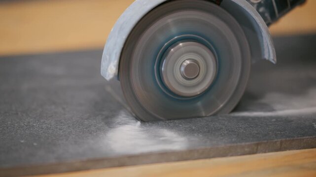 A person is cutting a ceramic tile with an electric grinder on a workbench during a home renovation project. Spinning diamond blade creates dust as it shapes the tile.