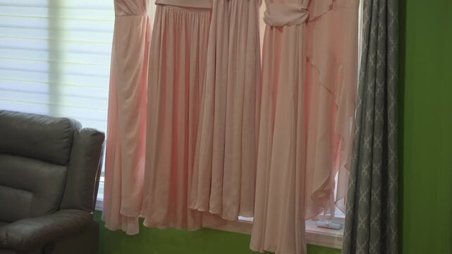A smooth upward tilt shot revealing four matching blush pink bridesmaid dresses hanging on a curtain rod in front of a window, showcasing different necklines and flowing fabrics.