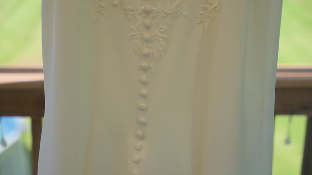 A smooth upward tilt shot of a white bridal dress, highlighting a row of covered buttons, intricate floral lace embroidery, and a sparkling jeweled belt against a soft green background.