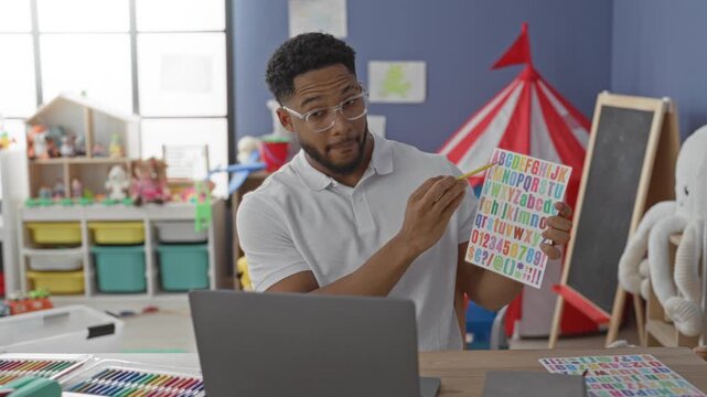 Man pointing pencil with hand at an alphabet card while seated at a laptop in a classroom building; focused teaching.