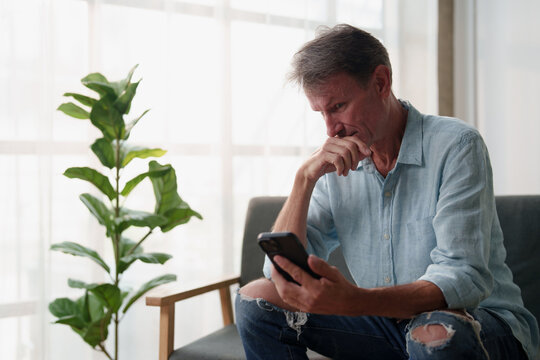 Mature man looking down at cell phone screen, hand on mouth, experiencing concern or bad news, contemplating a serious issue at home