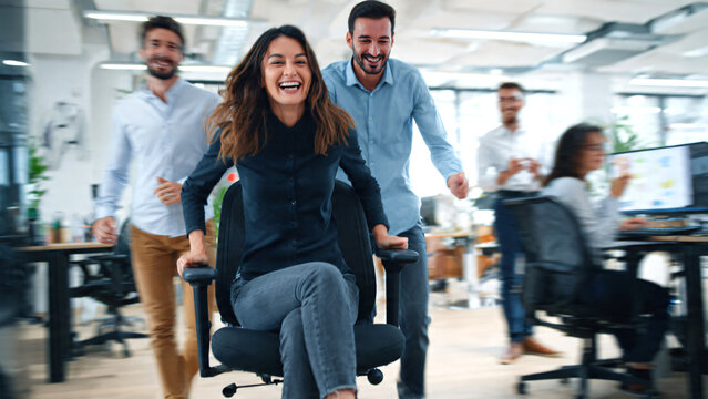 Happy colleagues celebrating success at workplace, two young coworkers playfully pushing smiling female CEO boss in office chair,  joyful teamwork, victory and positive corporate  Modern office.
