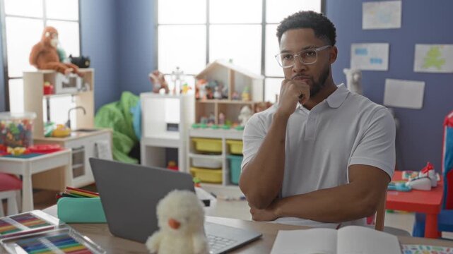 Man typing on laptop at a desk in a preschool classroom, hands on keyboard and open notebook; focused patience.