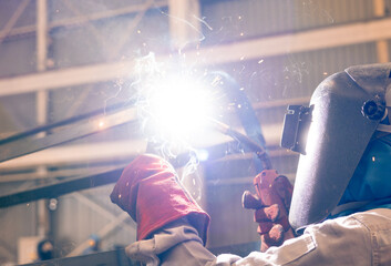 A worker stands in a workshop wearing safety gear while welding metal pieces together. Sparks fly from the welding tool as the task requires focus and precision © Happy Photo