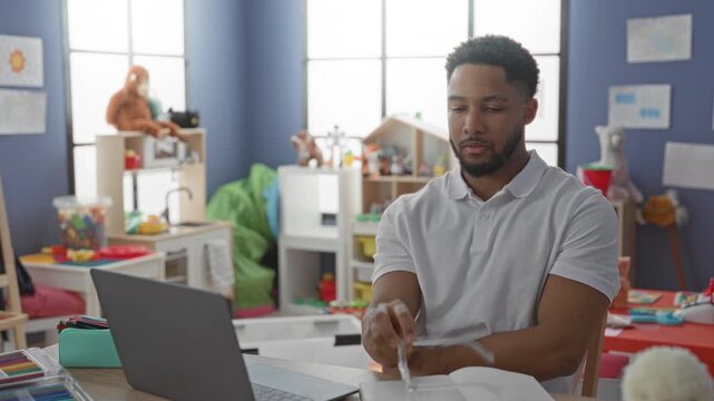 Man rubbing eyes at laptop amid scattered toys and open book in a colorful preschool classroom building, hand over face showing strain; fatigue stress.