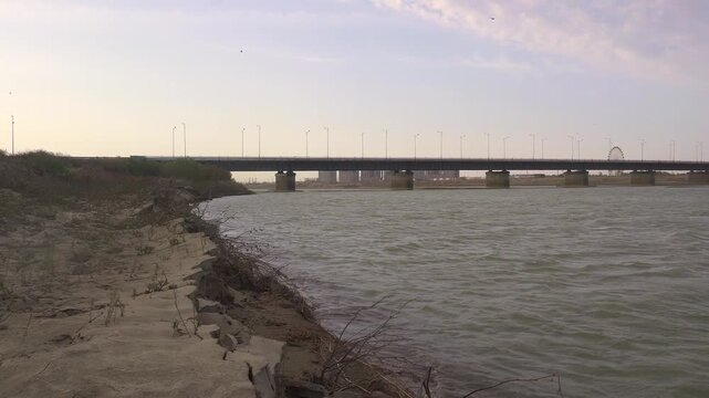 Sandy riverbank of the Syr Darya with gentle waves and a large highway bridge in the background, Kyzylorda, Kazakhstan.