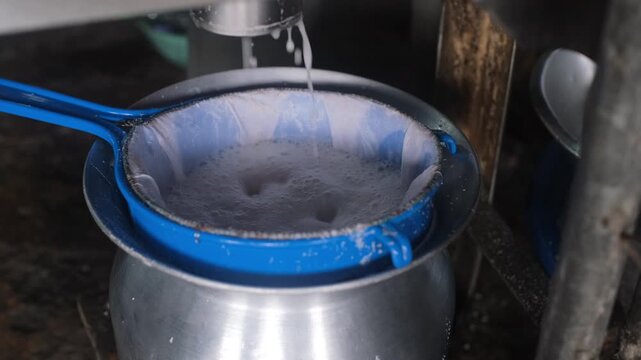 Grated coconut pressed and filtered through sieve with liquid coconut milk pouring into metal container on market stall. Traditional food preparation concept