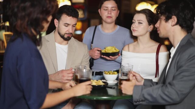  Group of male and female friends sit at table in bar enjoying conversation and drinking cold beer