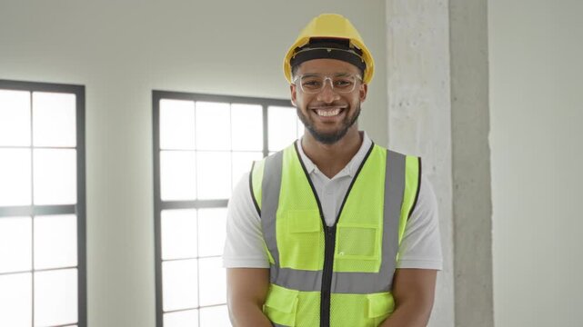 Man in hardhat and reflective vest smiling with face visible, standing by concrete pillar and windows in a building interior; confidence safety.