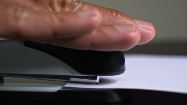 Side profile view of a hand pressing an office stapler down onto blank white paper