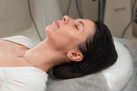 Close up of cosmetologist applying special facial gel on young woman before peeling procedure in beauty clinic. Skin preparation for professional cosmetology treatment. Skincare and aesthetic concept.