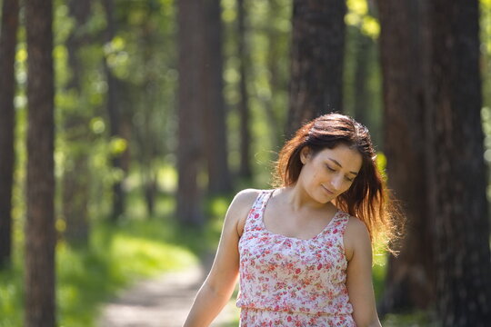 Portrait of a young woman in a park. A pretty woman stands on a path between pine trees, looking down, smiling mysteriously.