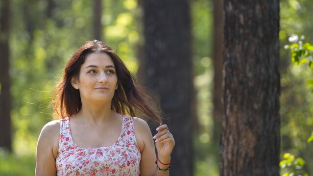 Portrait of a young woman in a park. A woman stands on a path between pine trees, lightly touching the ends of her hair. The background is beautifully blurred.