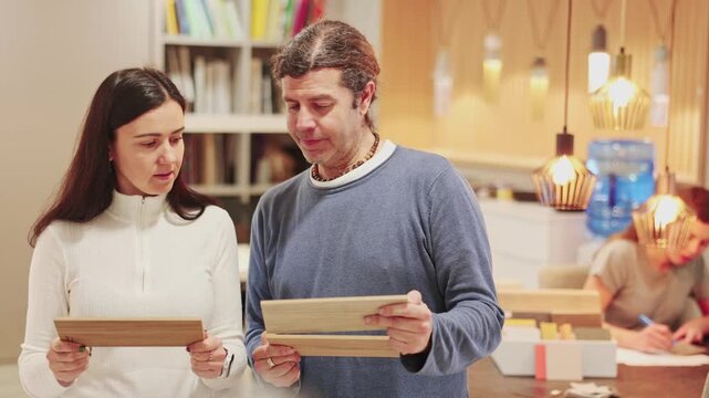 European married couple considering wood samples for apartment decoration and renovation, they choose laminate flooring. Buyers in a furniture salon discuss the quality of wooden materials. High