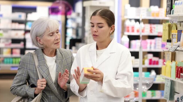 In pharmacy, girl apothecary holds package with therapeutic cream for sensitive skin in hands and talks with senior woman client about nuances of care. Colleague designs showcase in background
