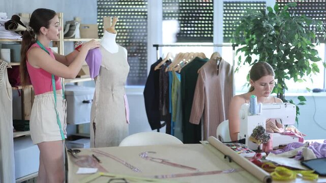 Two qualified dressmakers work in a sewing workshop against the backdrop of racks with fabrics and sewing accessories. Young women at a sewing workshop. High quality 4k footage