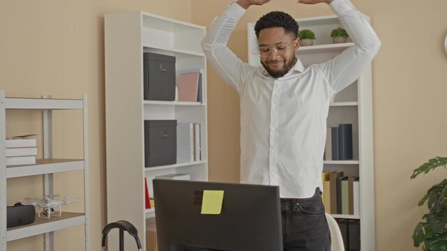 Man stretching arms above computer monitor while standing at desk in building, glasses on, bookshelf and clock visible; relaxed break.