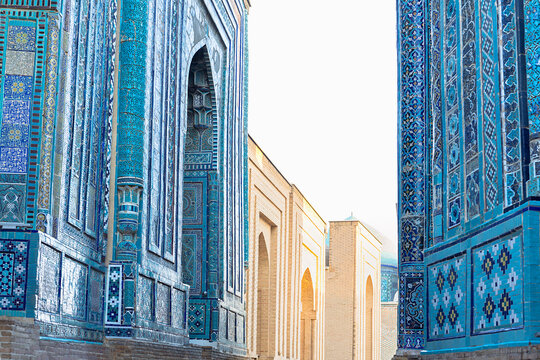 Shah-i-Zinda necropolis facades with intricate blue tilework in Samarkand. Ornate mausoleum walls line narrow passage bathed in daylight. Ideal for travel editorial, cultural heritage publications.