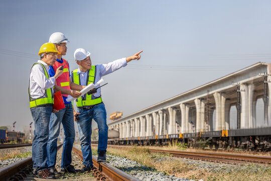 Group of asian railway engineers in safety gear discussing project with blueprint at construction site, Professional site manager and inspector pointing at infrastructure development near train tracks