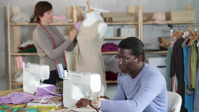 Experienced black middle-aged tailor stitching linen garment on sewing machine in workshop, while female colleague adjusting sample dress on mannequin