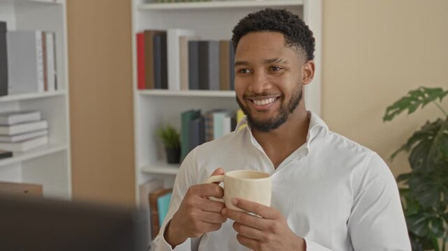 Man holding ceramic coffee mug with both hands in building while smiling at computer screen and wearing white shirt; calm productivity.