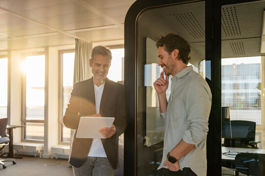 Two men having a business meeting in an office at sunset, using a tablet