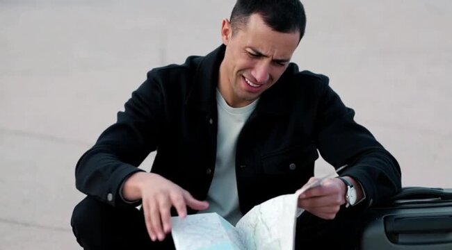 Young man reading map while sitting on suitcase near road, exploring travel routes and destinations during journey for adventure and navigation purpose