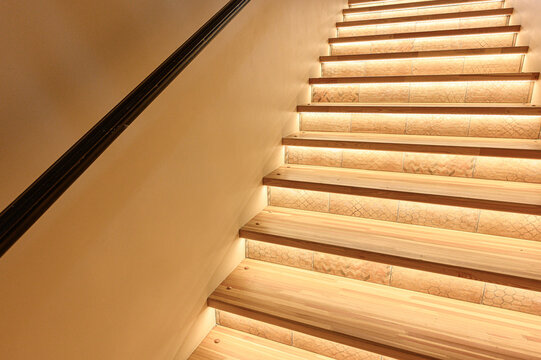 A modern staircase with wooden steps, built-in LED lighting under each step, light beige walls, and a dark handrail. Soft glow creates a warm, inviting atmosphere.
