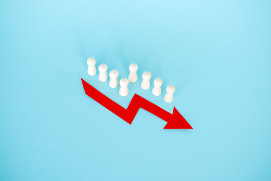 Line of wooden peg dolls stands behind red downward arrow on blue surface. Declining birth rate, demographic collapse trend, aging population crisis, Population shrinking statistics.