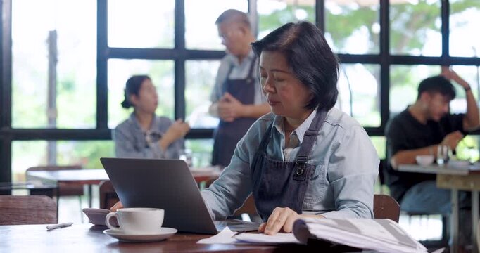 Asian senior female cafe owner in apron reviewing reports on laptop writing notes at table with coffee, teamwork management concept representing small family business showing responsibility dedication