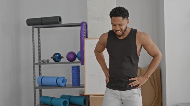Man rubbing forehead then hands on hips after workout among yoga mats and battle ropes in studio; breath recovery fatigue.
